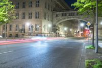 car lights streaking down an empty urban street at night with a large stone building and bridge