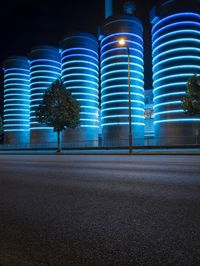 large cylindrical blue light at night near a building in a city with trees and bushes