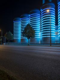 large cylindrical blue light at night near a building in a city with trees and bushes