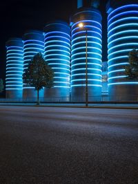 large cylindrical blue light at night near a building in a city with trees and bushes