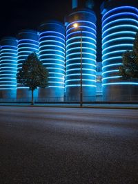 large cylindrical blue light at night near a building in a city with trees and bushes