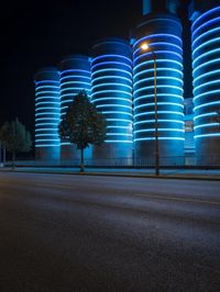 large cylindrical blue light at night near a building in a city with trees and bushes