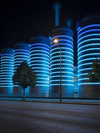 large cylindrical blue light at night near a building in a city with trees and bushes