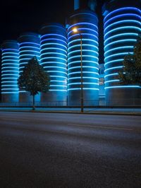 large cylindrical blue light at night near a building in a city with trees and bushes