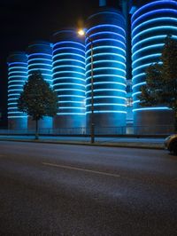 large cylindrical blue light at night near a building in a city with trees and bushes