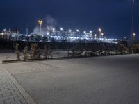 an empty street in front of a large industrial site lit up at night with many lights on it