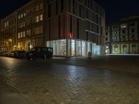 night time scene of a city street with parked cars and a glass building on the corner