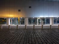 an empty sidewalk lit up at night next to a building with glass walls and doors