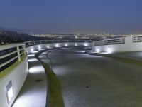 a circular walkway lit up with bright lights above a city at night time with urban buildings in the distance