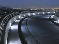 a circular walkway lit up with bright lights above a city at night time with urban buildings in the distance