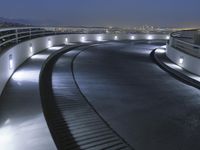 a circular walkway lit up with bright lights above a city at night time with urban buildings in the distance