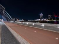 a deserted, empty road that runs alongside the bridge over the water at night in front of a large city skyline