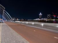 a deserted, empty road that runs alongside the bridge over the water at night in front of a large city skyline