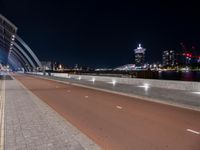 a deserted, empty road that runs alongside the bridge over the water at night in front of a large city skyline