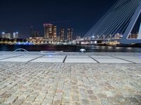 a bench next to the water near a bridge at night with buildings in the background