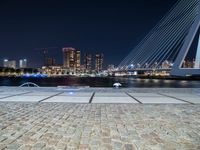 a bench next to the water near a bridge at night with buildings in the background