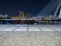 a bench next to the water near a bridge at night with buildings in the background