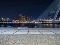 a bench next to the water near a bridge at night with buildings in the background