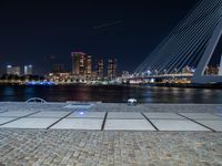 a bench next to the water near a bridge at night with buildings in the background