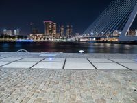 a bench next to the water near a bridge at night with buildings in the background