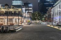 a city at night with an illuminated street and sidewalk area with benches and parked vehicles