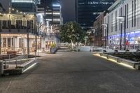 a city at night with an illuminated street and sidewalk area with benches and parked vehicles