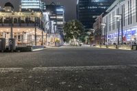 a city at night with an illuminated street and sidewalk area with benches and parked vehicles