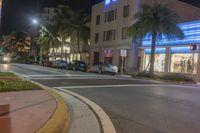 several cars parked on the sidewalk and a street corner at night with traffic lights at both end and palm trees at the sidewalk