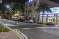 several cars parked on the sidewalk and a street corner at night with traffic lights at both end and palm trees at the sidewalk