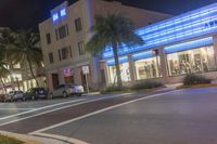 several cars parked on the sidewalk and a street corner at night with traffic lights at both end and palm trees at the sidewalk