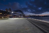 an empty plaza at dusk next to the ocean with a big bridge and city skyline in background