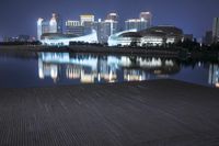 a pier at night with the lights reflecting on a water way and large buildings and tall structures in the distance
