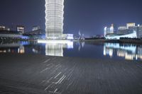 a pier at night with the lights reflecting on a water way and large buildings and tall structures in the distance