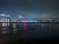 a city lit up on the waterfront at night with some lights reflecting in water and clouds