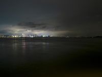 a city lit up on the waterfront at night with some lights reflecting in water and clouds