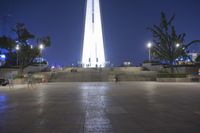 people walking along a plaza near a tall obelisk in the night, with an illuminated streetlight shining on it