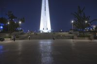 people walking along a plaza near a tall obelisk in the night, with an illuminated streetlight shining on it