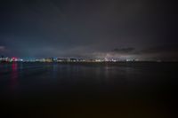 dark and cloudy night sky over city lights from pier in foreground with reflecting water, buildings with red lights in distance