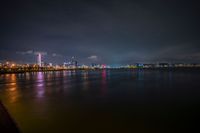 dark and cloudy night sky over city lights from pier in foreground with reflecting water, buildings with red lights in distance