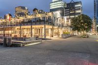 a group of buildings at the beach next to a bench on the sidewalk, with lights lit up and overlooking a city skyline