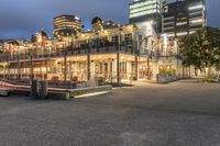 a group of buildings at the beach next to a bench on the sidewalk, with lights lit up and overlooking a city skyline