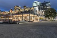 a group of buildings at the beach next to a bench on the sidewalk, with lights lit up and overlooking a city skyline