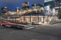 a group of buildings at the beach next to a bench on the sidewalk, with lights lit up and overlooking a city skyline