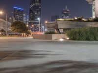 a long cement bench next to a walkway near tall buildings at night with lights on