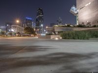 a long cement bench next to a walkway near tall buildings at night with lights on