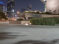 a long cement bench next to a walkway near tall buildings at night with lights on