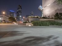 a long cement bench next to a walkway near tall buildings at night with lights on