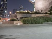 a long cement bench next to a walkway near tall buildings at night with lights on