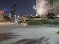 a long cement bench next to a walkway near tall buildings at night with lights on