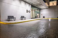 empty benches lined up outside a building at night time on a rainy day with the sidewalk painted yellow